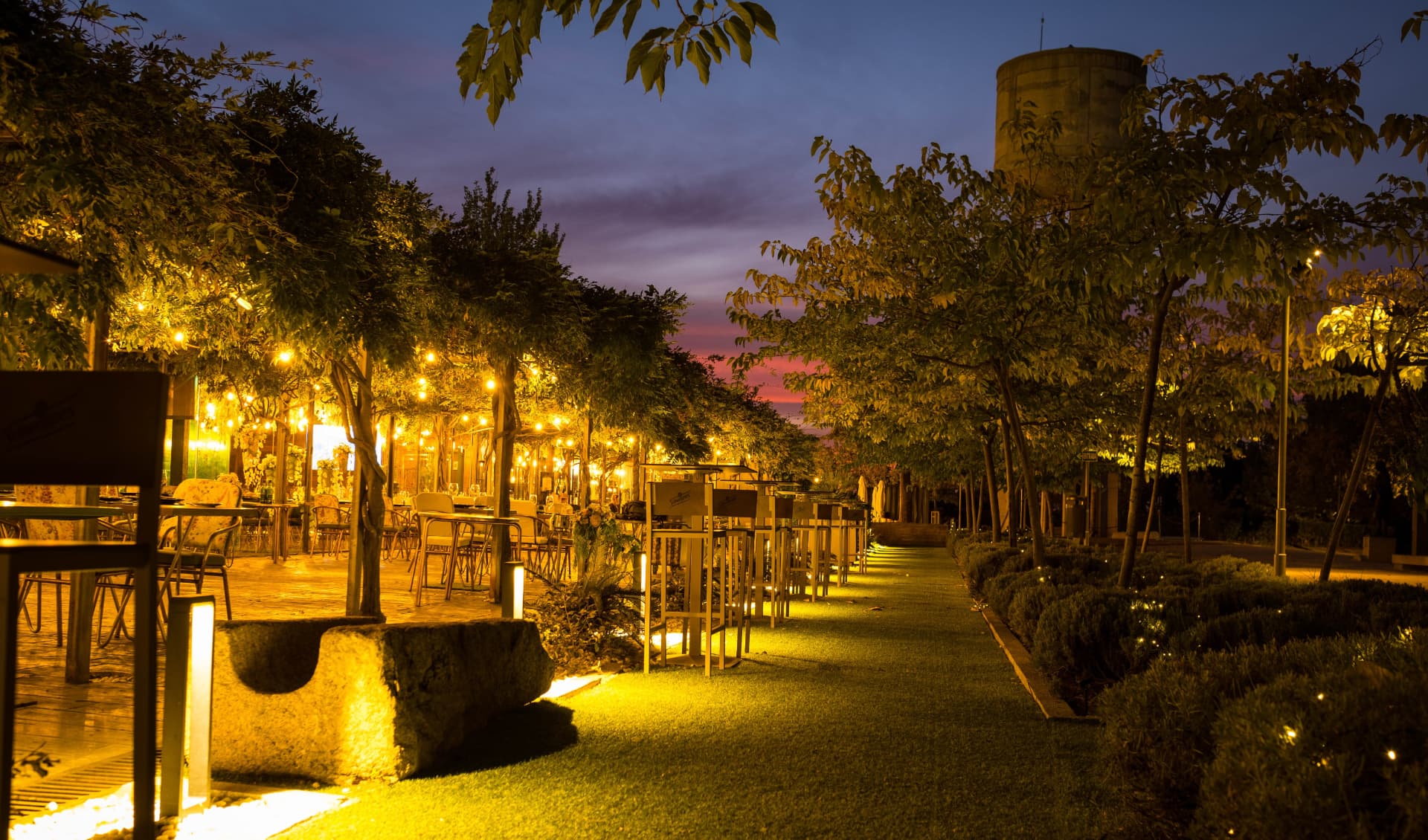 Paseo del jardín de El Jardín de Nora iluminado al anochecer, con árboles, guirnaldas y el torreón al fondo